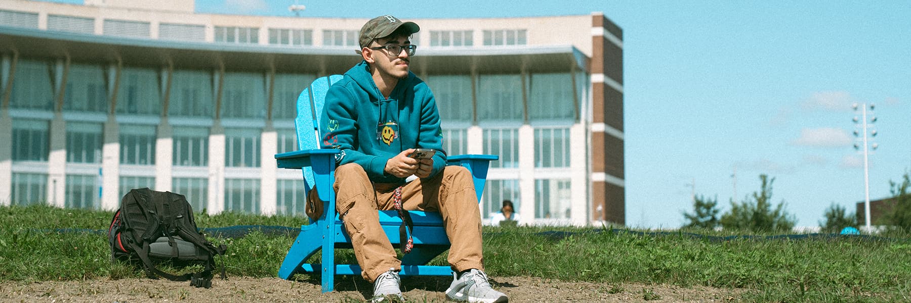 Student sits in blue chair on campus center lawn with cellphone in hand.