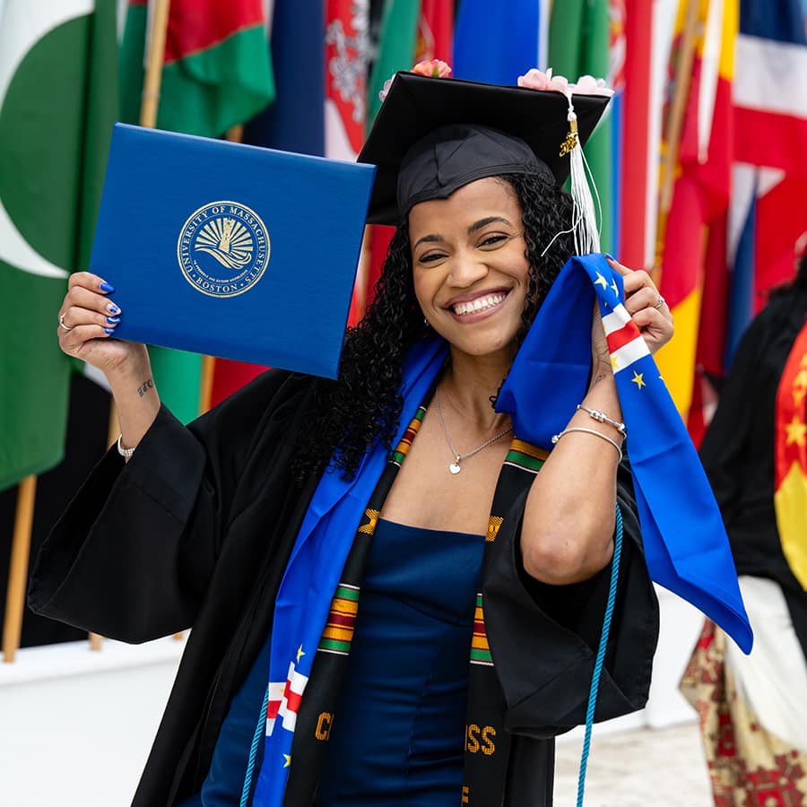 Student in cap and gown holding up their diploma in front of row of flags.