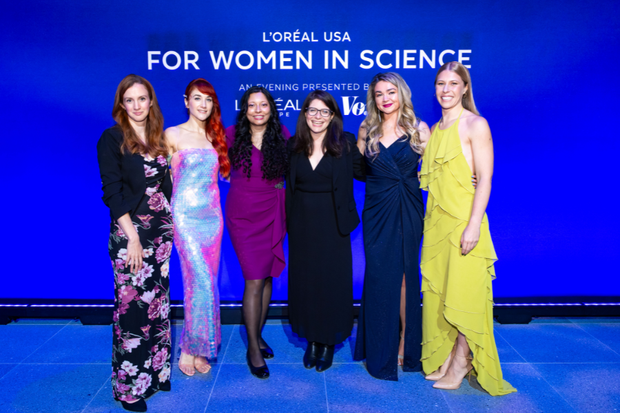 Six women pose on a stage in front of a bright blue backdrop reading “L’Oréal USA For Women in Science.
