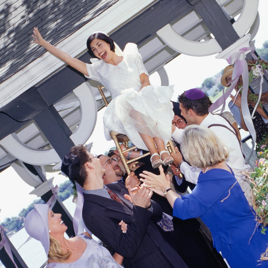Men carrying bride on chair at Jewish wedding ceremony while people around clap in celebration