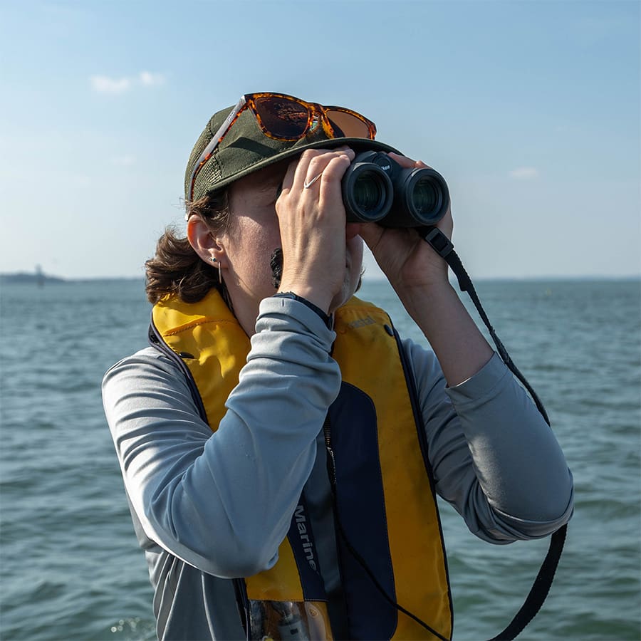 Student researcher on a boat looking through binoculars.